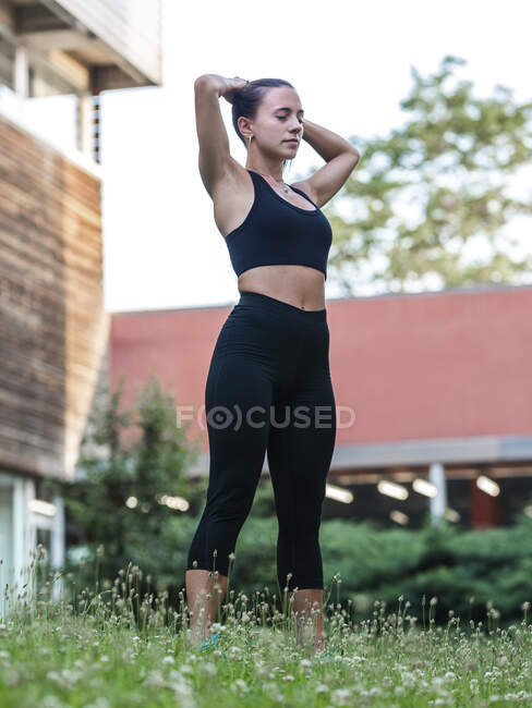 Young sportswoman with closed eyes doing ponytail during fitness workout on lawn — Stock Photo