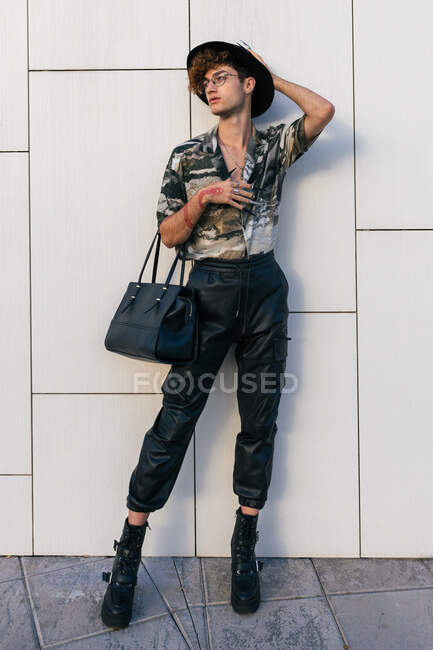 Молодой тщеславный мужчина в стильной одежде с дамской сумочкой, стоящей на черепичной стене, отводя взгляд — стоковое фото