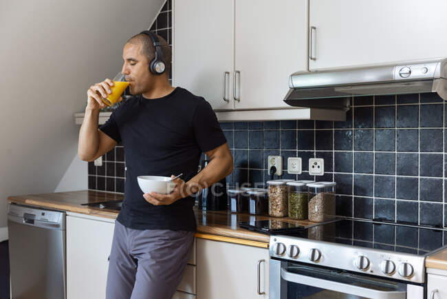 Maschio in cuffia ascoltare musica e bere succo d'arancia fresco mentre fa colazione al mattino a casa e in piedi vicino al bancone in cucina — Foto stock