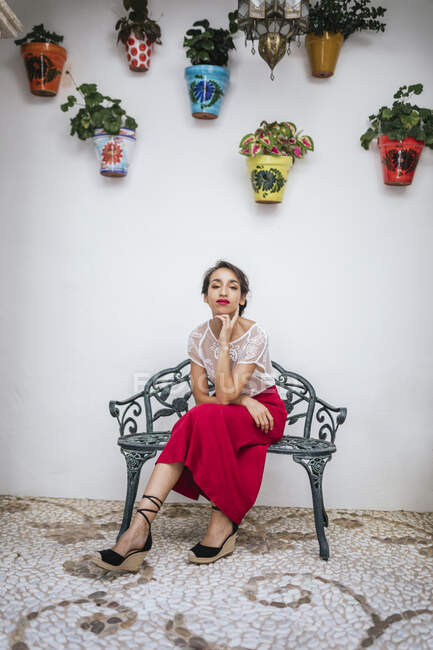 Anmutige ethnische Frau in rotem Rock und Lippen sitzt im Sommer auf einem Hocker im Innenhof des Hauses, während sie den Hals berührt und in die Kamera blickt — Stockfoto