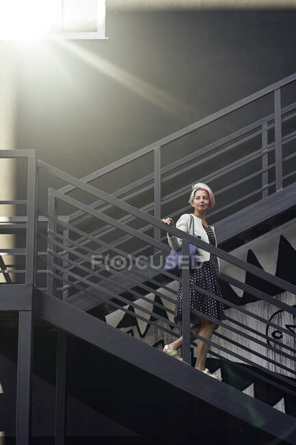 Délicieuse femelle en robe debout sur un escalier noir éclairé par la lumière du soleil et regardant loin — Photo de stock