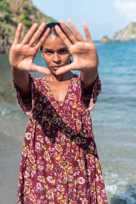 Turista femenina étnica seria en vestido de sol demostrando gesto de triángulo mientras mira a la cámara en la playa del océano - foto de stock