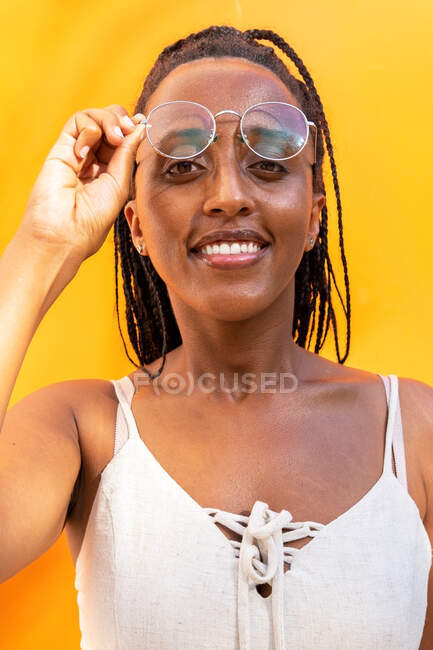 Mujer afroamericana positiva con gafas trenzadas tocando el pelo y mirando a la cámara sobre fondo amarillo en Barcelona en verano - foto de stock