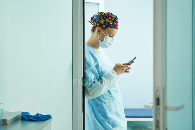 Vista lateral del médico femenino en uniforme médico y mensajes de texto de máscara estéril en el teléfono celular en la clínica - foto de stock