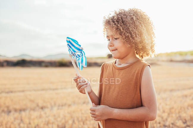 Vista lateral da criança de cabelos encaracolados em pé no prado seco e brincando com moinho de vento de brinquedo no verão — Fotografia de Stock