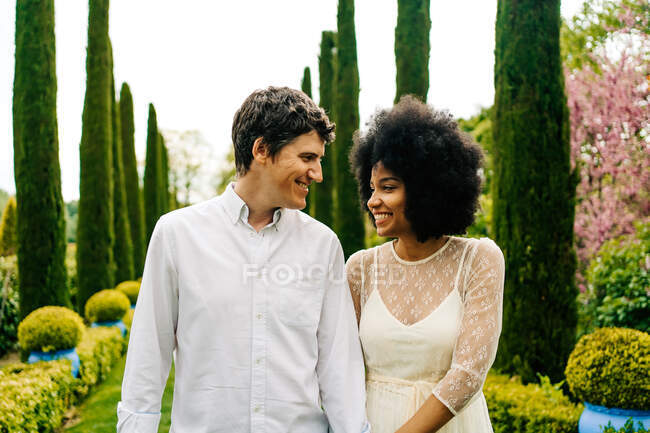 Fröhliches multiethnisches Paar, das sich Händchen hält und im Garten spazieren geht, während es einander ansieht — Stockfoto