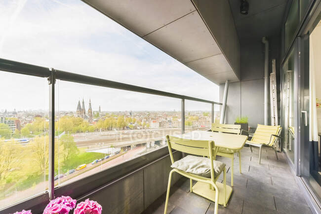 Mesa con sillas situada en el amplio balcón del edificio residencial contemporáneo con cerca de cristal que mira paisaje urbano en el día de verano - foto de stock
