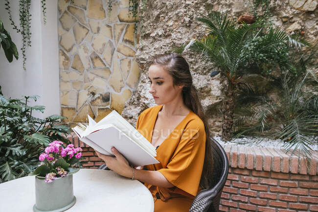 Side view of serene tender female reading interesting book while sitting at table on summer terrace of house — Stock Photo