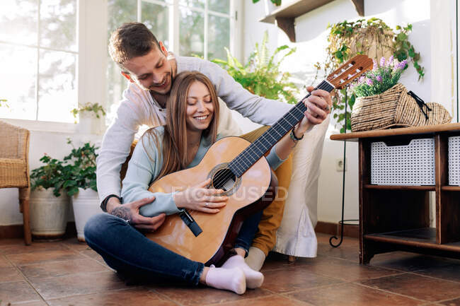 Веселый татуированный мужчина-музыкант играет на гитаре рядом с веселым татуированным мужчиной-музыкантом, глядя друг на друга в кресле в комнате — стоковое фото