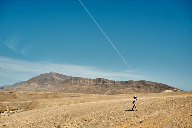 Мандрівниця - самотня жінка, яка в сонячний день ходить у сухій долині поблизу гірського пасма проти блакитного неба у Фуертевентурі (Іспанія). — стокове фото