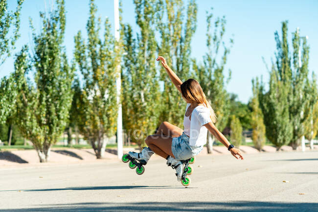 Vista lateral del ajuste femenino balanceo en patines y mostrando un truco de una pierna con ruedas mientras está en la carretera en la ciudad en verano - foto de stock