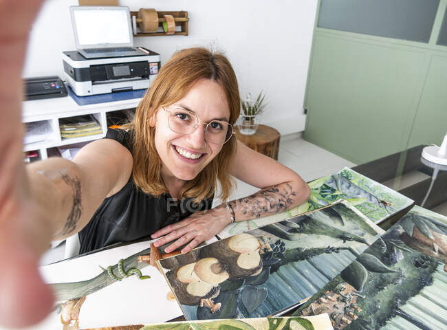 Cheerful female designer sitting at table with art supplies and taking self shot on smartphone in workspace — Stock Photo