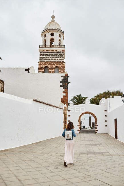 Vista trasera de una mujer anónima con mochila caminando sobre pavimento contra casas blancas y cielo gris nublado en la calle de la ciudad en Fuerteventura, España - foto de stock