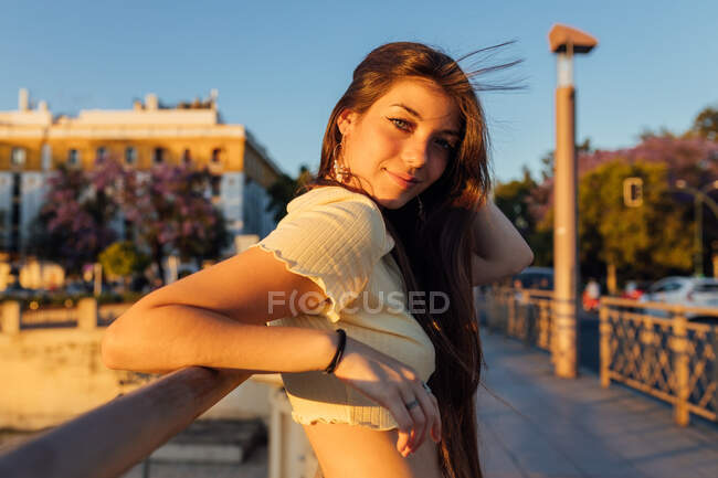 Side view of young gentle female with flying hair looking at camera against fence on bridge in soft sunlight — Stock Photo