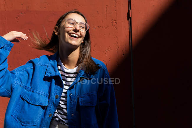 Femme positive en tenue élégante jetant les cheveux et regardant loin sur le fond du mur de bâtiment par une journée ensoleillée dans la rue de la ville — Photo de stock