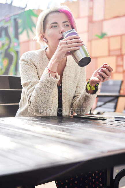 Low angle of pensive alternative female with dyed hair drinking hot beverage from eco friendly cup while sitting at table in street cafe and looking away — Stock Photo