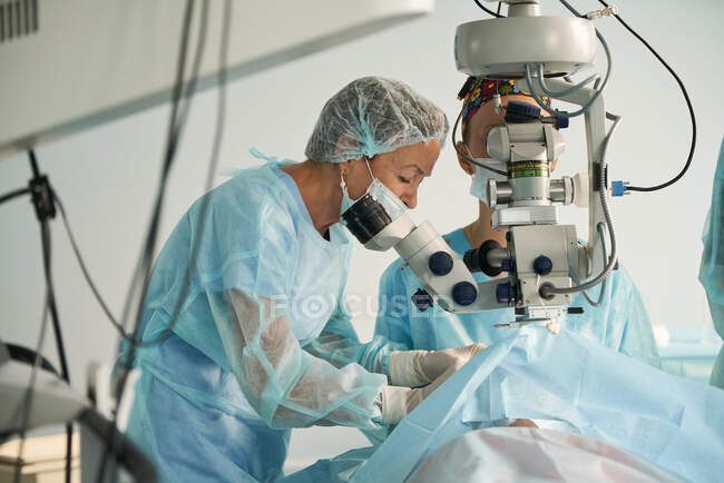 Dos mujeres adultas doctora en máscara estéril y gorra médica ornamental mirando a través del microscopio quirúrgico contra compañeros de cultivo en el hospital - foto de stock
