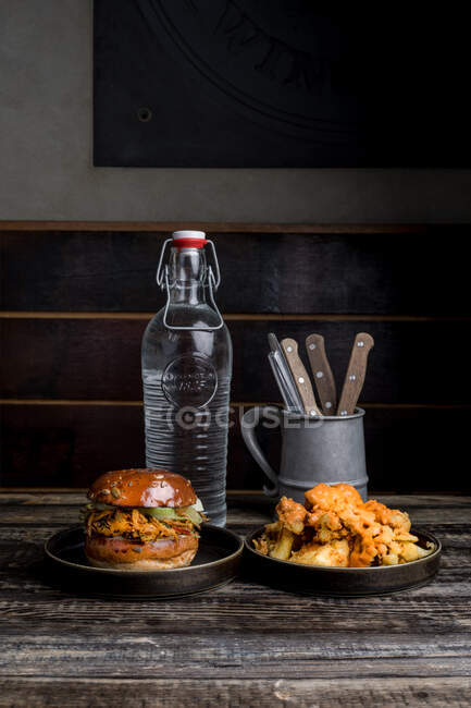Hambúrguer saboroso com caixas fritas colocadas perto da placa com frango crocante na mesa de madeira contra garrafa de água e utensílios de mesa no restaurante — Fotografia de Stock