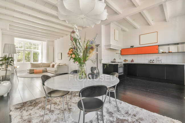 Diseño interior de comedor con mesa redonda y sillas colocadas en alfombra en amplio apartamento con cocina abierta y paredes blancas y techo en estilo loft moderno - foto de stock