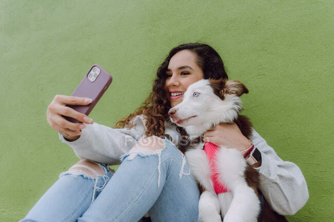 Zufriedene Besitzerin macht Selbstporträt mit Border Collie Hund auf Smartphone, während sie in der Nähe der grünen Mauer in der Stadtstraße sitzt — Stockfoto