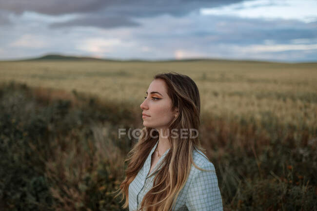 Vista lateral de la joven hembra consciente mirando hacia otro lado en el camino cerca del prado bajo el cielo nublado por la noche en el campo - foto de stock
