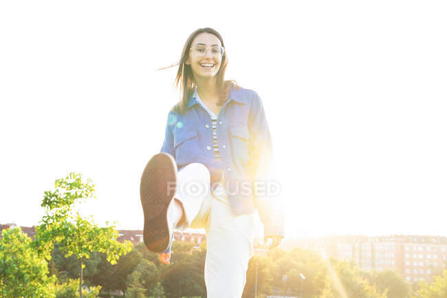 Baixo ângulo de elegante positivo feminino chutando perna para câmera no fundo da cidade em volta iluminado — Fotografia de Stock