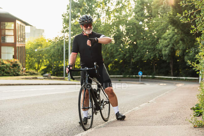 Middle aged ethnic sportsman on bicycle watching blood pressure on wearable tracker during workout in summer park — Stock Photo