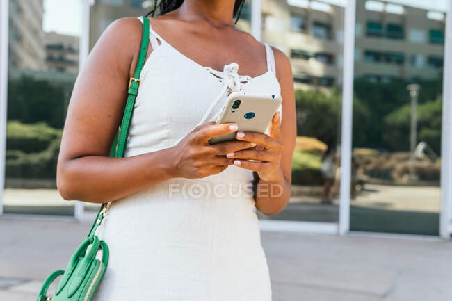 Bajo ángulo de mujer afroamericana anónima con brads mensajería en las redes sociales a través del teléfono móvil mientras está de pie en la calle con edificios antiguos en Barcelona - foto de stock