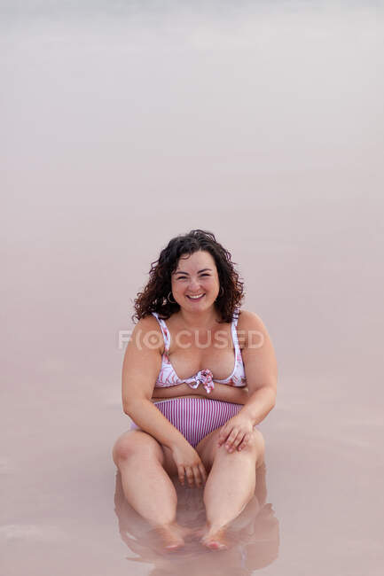 Deleitada hembra curvilínea en bikini sentada en agua de estanque rosa en verano y mirando a la cámara - foto de stock