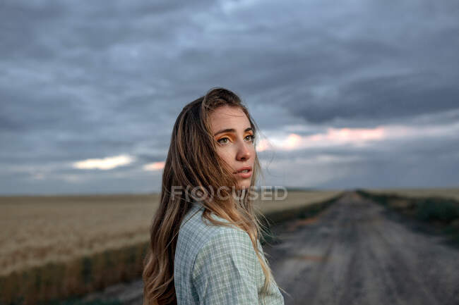 Vista lateral de la joven hembra consciente mirando hacia otro lado en el camino cerca del prado bajo el cielo nublado por la noche en el campo - foto de stock