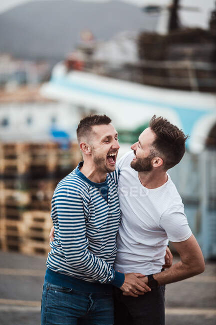 Содержание пара гомосексуальных мужчин в футболках обнимаются, разговаривая и глядя друг на друга в городе — стоковое фото