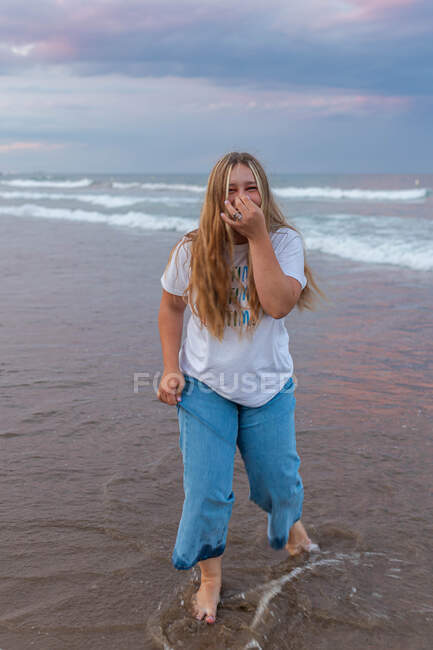 Delighted young curvy female standing barefoot on wet beach near sea and having fun while looking at camera and touching face — Stock Photo