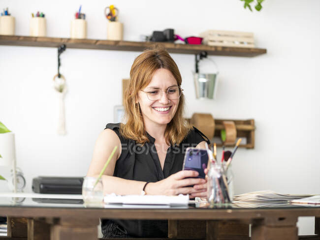 Designer feminino alegre usando smartphone enquanto se senta na mesa e trabalha no espaço de trabalho criativo — Fotografia de Stock