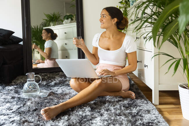 Созерцательная этническая женщина с нетбуком и стаканом воды, отдыхающая на мягком ковре, глядя в сторону от зеркала в доме — стоковое фото