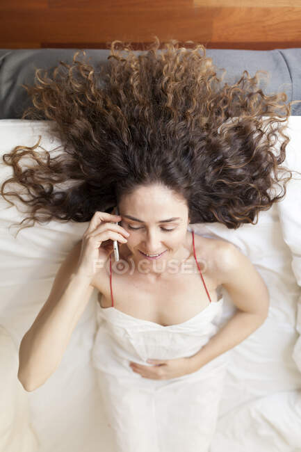 Вид сверху деловой женщины с кудрявыми волосами, лежащей в кровати и разговаривающей по телефону — стоковое фото