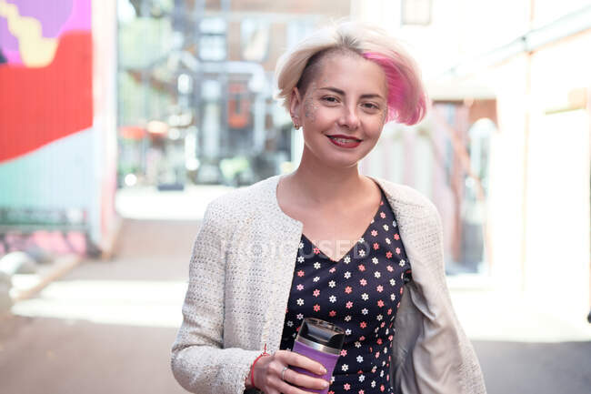 Вид спереди радостной альтернативной женщины с окрашенными волосами и курткой, стоящей на улице с чашкой горячего напитка и смотрящей в камеру — стоковое фото