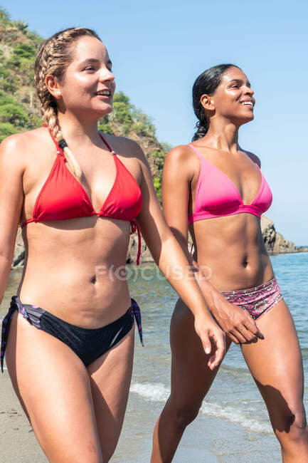 From below content multiethnic female travelers in swimwear speaking on ocean beach against rocks during summer journey in sunlight — Stock Photo