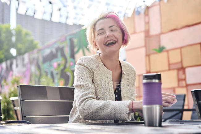 Low angle of happy alternative female with dyed hair having breakfast while sitting at table in street cafe with closed eyes — Stock Photo