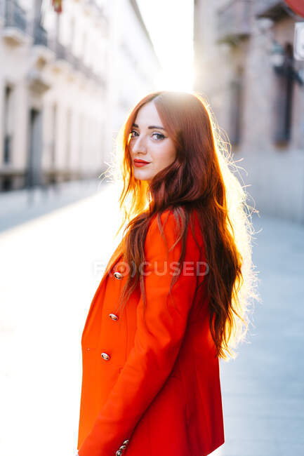 Vue latérale de la femme élégante avec des cheveux roux et en costume orange vif marchant dans la rue de la ville en regardant la caméra — Photo de stock