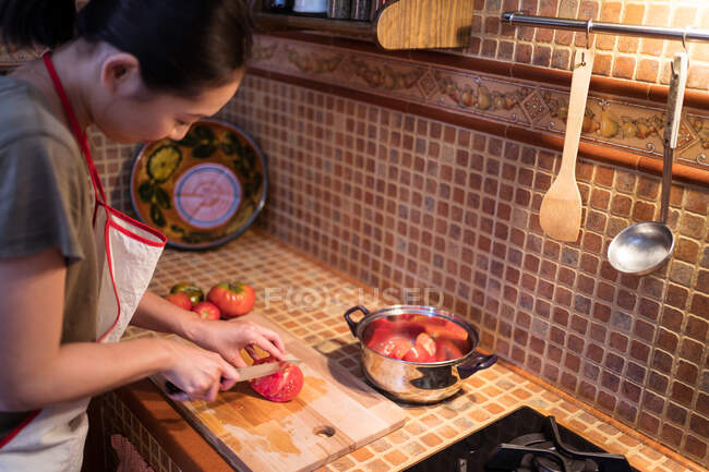 Сверху вид этнической женщины в фартуке режет спелые помидоры на доске во время приготовления обеда на кухне дома — стоковое фото