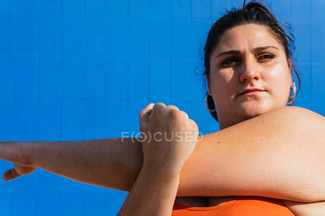 Entschlossene mollige ethnische Sportlerin beim Training, während sie an einem sonnigen Tag vor blauem Hintergrund wegschaut — Stockfoto