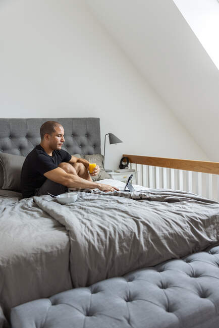 Seitenansicht eines Mannes, der mit einem Glas Orangensaft und einem Tablet auf dem Bett sitzt, während er morgens zu Hause frühstückt — Stockfoto