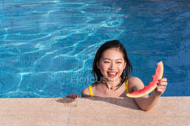 Mulher étnica alegre na piscina com fatia de melancia enquanto olha para longe no dia ensolarado no verão — Fotografia de Stock