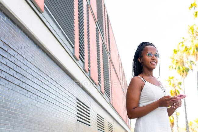 Angolo basso di donna afroamericana sorridente con messaggi brads sui social media tramite telefono cellulare mentre si trova in strada con vecchi edifici a Barcellona — Foto stock
