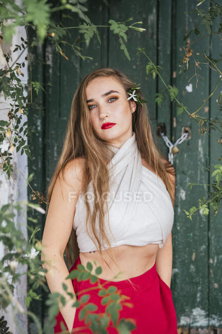 Mulher graciosa com lábios vermelhos e em roupa de verão desfrutando de aroma de flores aromáticas crescendo no pátio da casa olhando para a câmera — Fotografia de Stock
