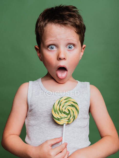 Deleitado niño preadolescente con caramelo remolino con la lengua hacia fuera mientras mira a la cámara en el fondo verde en el estudio — Stock Photo