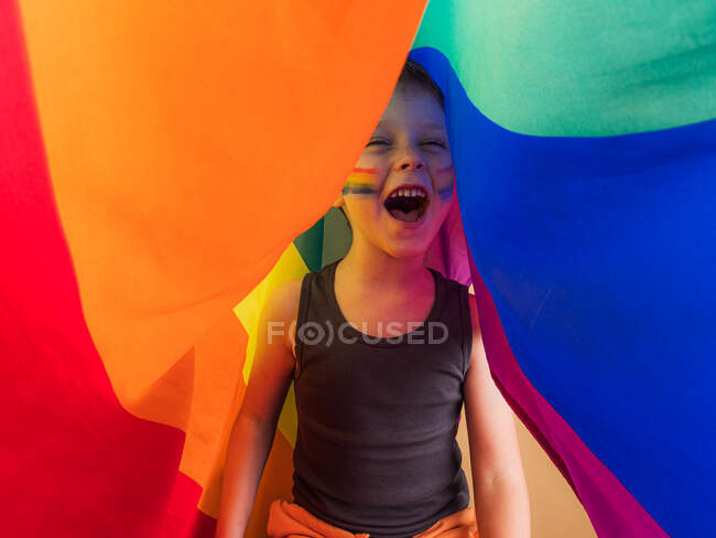 Positives Kind im Unterhemd mit geschminkten Wangen und offenem Mund, das schreit, während es sich unter LGBTQ-Flagge nach vorne freut — Stockfoto
