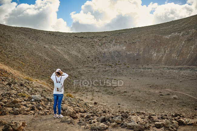 Visão traseira da mulher anônima em roupas casuais tocando a cabeça e olhando para a cratera árida com encostas pedregosas em Fuerteventura, Espanha — Fotografia de Stock