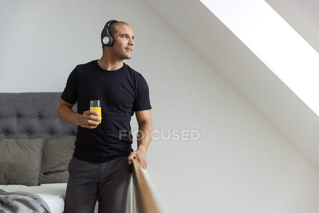 Вид збоку спокійного чоловіка з навушниками, що слухають музику, що стоїть біля ліжка після пробудження в спальні і насолоджується ранком під час пиття апельсинового соку — стокове фото