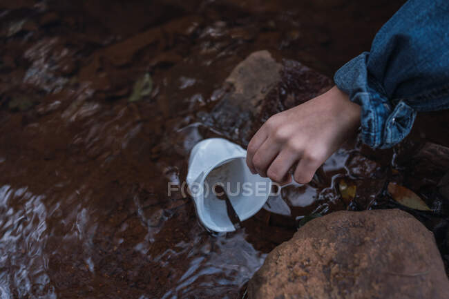 From above of crop anonymous person picking up disposable glass from shallow river with stones in daytime — Stock Photo
